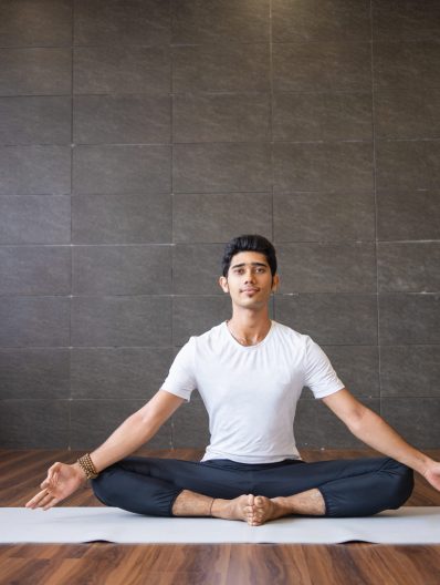 Indian yogi sitting and holding feet together in gym. Man looking at camera, keeping hands in mudra gesture and practicing yoga. Yogi concept. Front view.